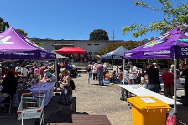 Outdoor community event with people mingling under colourful tents, tables with goods on a sunny day. Trees and a building are visible in the background.