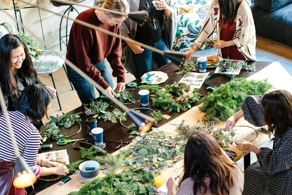 Group of people each crafting their Christmas wreath with natural foliage on table.