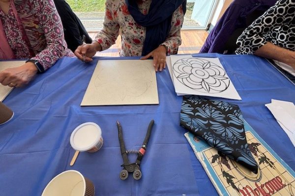 Three people sit at a table covered with a blue cloth, working on mosaics. Art supplies, a cup, a tool, and fabric are on the table.
