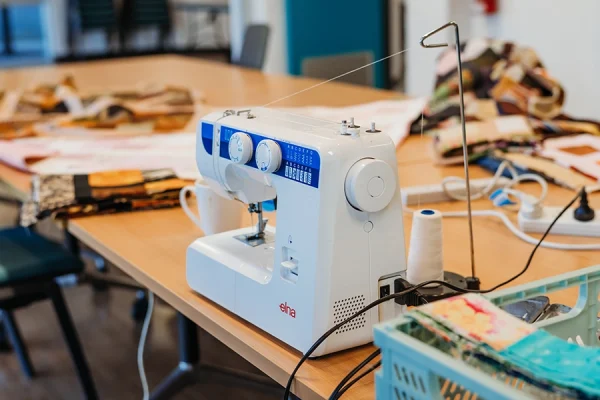 A white sewing machine sits on a wooden table surrounded by colourful fabric pieces and sewing supplies in a well-lit room.