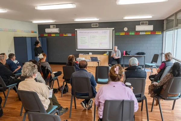 A group of people sit in a classroom arranged in a semi-circle, facing a presenter and a projection screen displaying slides at the front of the room. The atmosphere is casual and attentive.