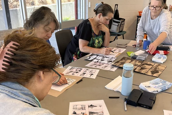 Four women sit around a table working on art projects with paper, pens, and reference images. One woman is leaning over to reach something, while the others are focused on drawing or colouring.