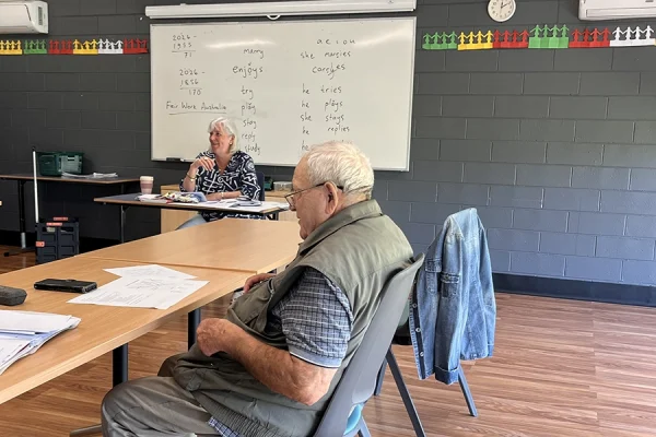 Two adults sit in a classroom; one is at a desk at the front of the room, while the other sits at a table with papers. A whiteboard with writing is visible behind them on a grey brick wall.