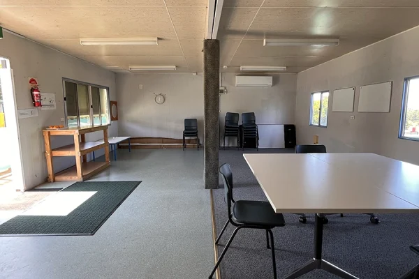 A simple, empty room with tables and chairs, white walls, fluorescent lighting, a stack of chairs in the corner, and plenty of natural light from the windows. A clock is on the wall, and a door is partially open.