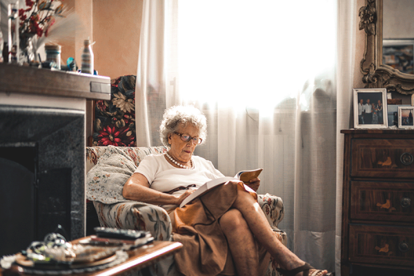 An elderly woman with glasses sits in an armchair by a window, reading a book in a cosy living room.