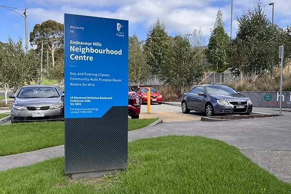 A blue sign for the Endeavour Hills Neighbourhood Centre stands by a car park with several parked cars, surrounded by green grass, trees, and a partly cloudy sky in the background.
