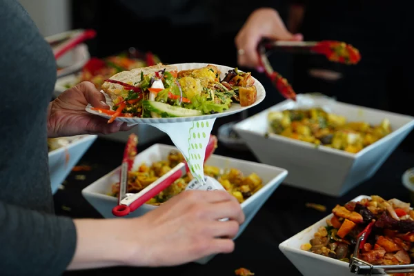A person holds a paper plate filled with salad and various colourful foods, serving themselves from trays of assorted dishes at a buffet-style Harmony Day event. Other hands with tongs are also reaching for food.
