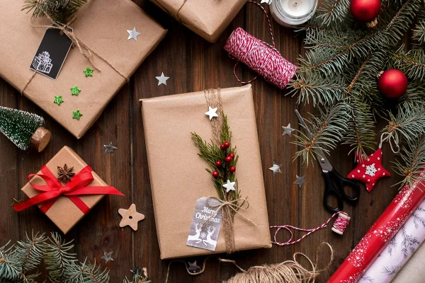 Christmas gifts wrapped in brown paper with festive decorations, red ornaments, pine branches, string, scissors, and wrapping paper rolls on a wooden table.