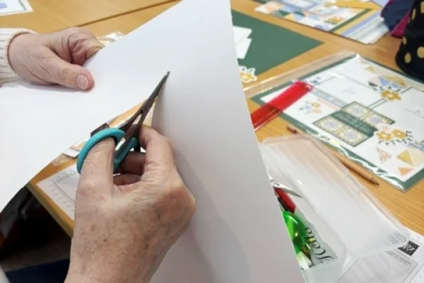 A person uses scissors to cut a sheet of white paper at a table covered with craft supplies, including rulers, patterned paper, and envelopes.