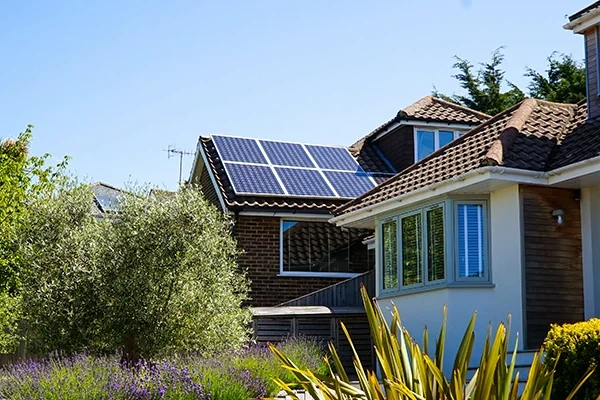 A suburban house with solar panels installed on the roof, surrounded by greenery and shrubs, under a clear blue sky.
